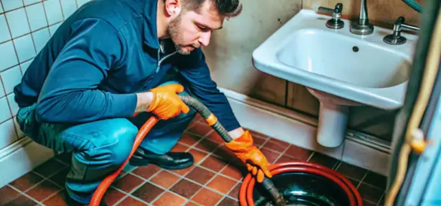 Plumber in blue coveralls crouching in a tiled bathroom and feeding an orange drain cable into a floor drain