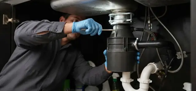 Plumber in blue gloves working on a garbage disposal under a kitchen sink
