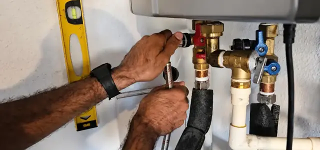 Hands tightening valves under a wall-mounted tankless water heater with a level on the pipe